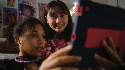 Asian teen lies on bed and watches content with friend. African American girl sits at floor, surfs internet using tablet. Multicultural girls spend leisure time together at home. Friends relationship.