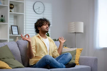 Tuinposter An Indian man sitting in the lotus pose on a couch, meditating with eyes closed in a peaceful living room setting. © Liubomir