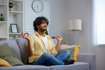 Peaceful Indian man with curly hair meditating in lotus pose on a sofa at home, exhibiting calmness...