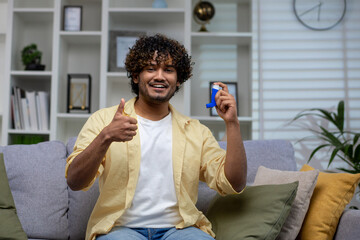Adult male in a casual outfit using an asthma inhaler while giving a thumbs up, seated comfortably on a couch at home.