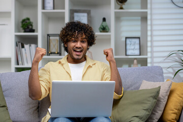 An overjoyed individual sitting on a couch with fists raised in victory, looking at a laptop screen...