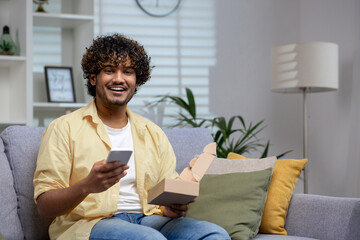A casual man sits comfortably on a couch at home, looking at his smartphone while holding an...