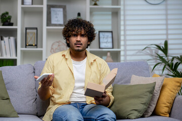 Perplexed Indian man sitting on couch with phone and open box, potentially facing online shopping scam. - obrazy, fototapety, plakaty