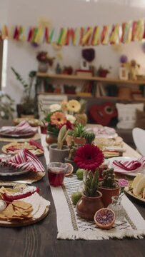 Close-up vertical no people footage of dinner table with tasty traditional Mexican food and snacks, for celebrating Cinco de Mayo - nachos, tacos, chalupa, salsa, guacamole, salad, flowers, cacti