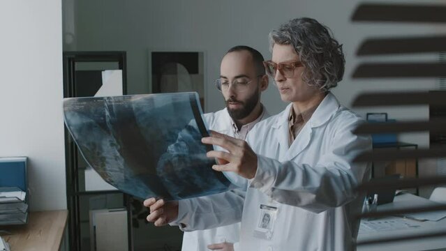 Medium shot of Caucasian female doctor in white coat and Arab male clinician standing in office, looking at xray of lungs on film, pointing to abnormalities, discussing diagnosis, treatment options