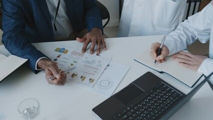 Medium close-up shot of hands of two unrecognizable male medical executive and doctor sitting at table during meeting, discussing slide with financial performance metrics, and improvements - Powered by Adobe