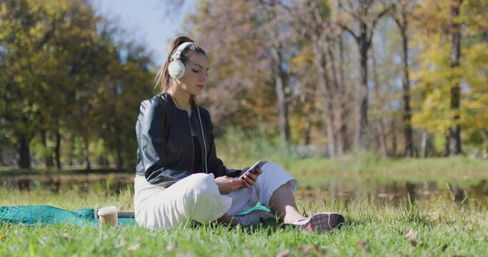 Beautiful girl sitting on a meadow in the park. Fashionable young lady with white headphones scrolling social media applications on her phone.