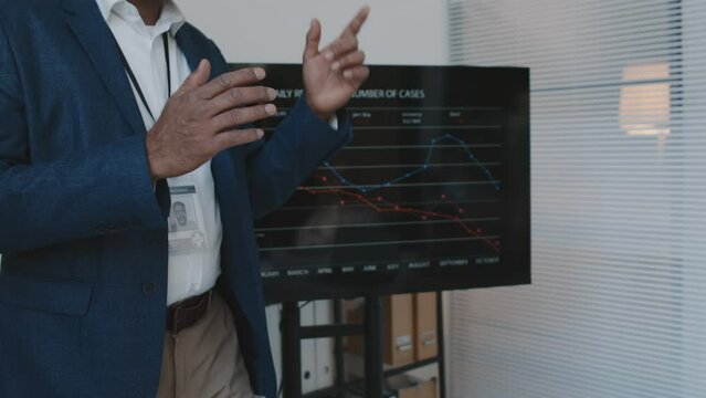 Medium shot of midsection of unrecognizable dark-skinned male CEO in suit standing by screen with presentation during meeting, gesticulating, explaining staff ways to improve hospital profitability