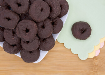 Cropped angle view of a square plate with round chocolate donuts on a light wood table, one donut isolated on green paper napkin with pink and yellow napkins below. Scalloped edges on napkins.