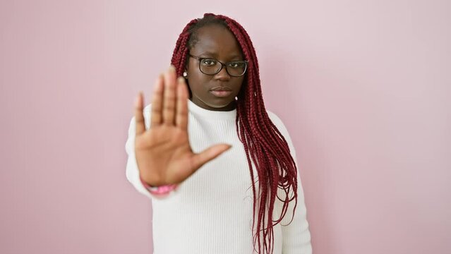 A confident african woman with braids wearing glasses stands against a pink background, gesturing 'stop' with her hand.