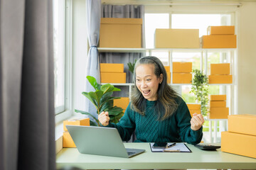 Starting a Small Business SME Excited elderly female business owner uses her smartphone or laptop to receive and check online orders to prepare boxes of products.