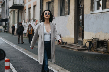 Trendy young woman in sunglasses striding with confidence through an urban setting, showcasing city...