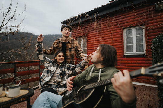 A group of friends gather on a wooden porch, playing guitar and enjoying the mountain view from their rustic cabin getaway. - Powered by Adobe