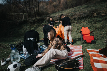 Group of friends setting up camp in nature, relaxing with a guitar on colorful blankets and preparing a tent. - Powered by Adobe