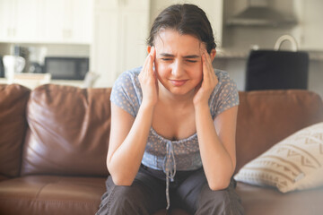 Young Woman Experiencing a Headache at Home. A teenage girl sits on a leather couch, hands pressed...