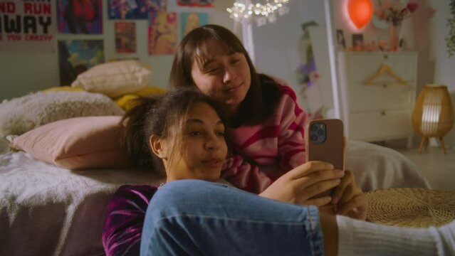 African American girl sits at floor and surfs internet using phone. Asian teenager lies on bed and watches at phone. Two multi ethnic girls together takes a picture using phone. Friends relationship.
