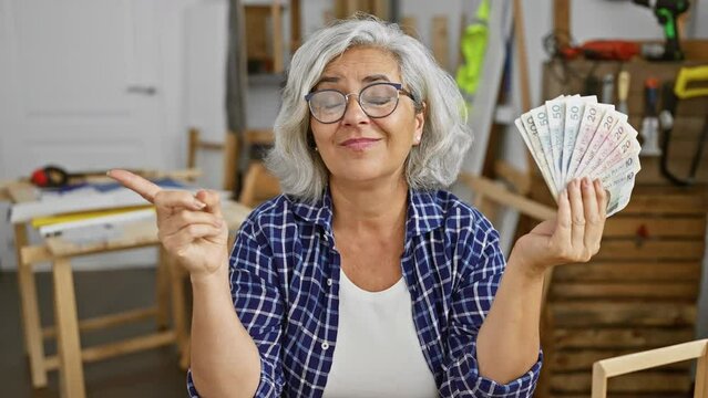 Cheerful middle-aged woman with grey hair joyfully clutching polish zloty banknotes in carpentry workshop, pointing aside with a friendly smile