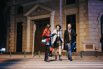 Three young business adults crossing the street at night, laughing and enjoying a city vibe, showcasing friendship and urban life. - obrazy, fototapety, plakaty