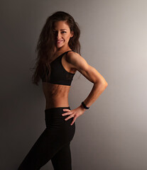 Sport smiling muscular woman posing in black sport bra with long brown hair standing on dark shadow studio background. Profile body view portrait. Sporty - 771141607