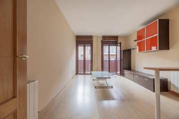 Living room of a loft-type house with an open kitchen, a small table wing with a leg, light wooden floors and two red aluminum balconies facing the street.