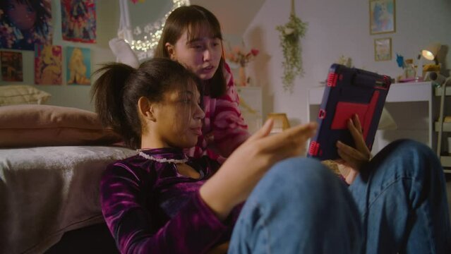African American girl sits at floor, surfs internet using tablet. Asian teen lies on bed and watches content with friend. Happy diverse girls together spend leisure time at home. Friends relationship.