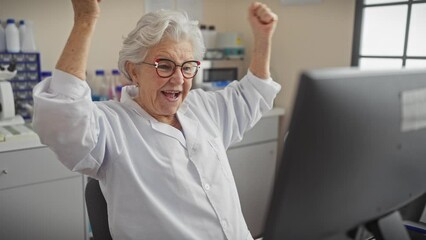 Excited grey-haired senior woman in scientist uniform screaming in joy, celebrating victory with raised arm at lab, full of triumph and exuberance!