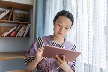 An Asian woman is studying diligently on a tablet in the living room desk at home