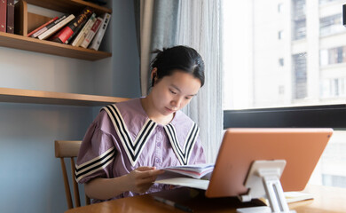 An Asian woman is studying diligently on a tablet in the living room desk at home