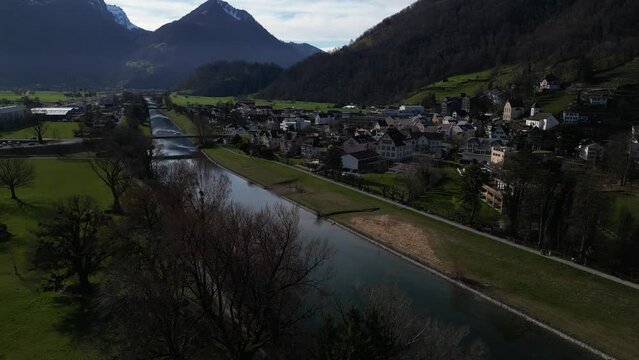 Drone clip over calm river passing traditional Alpine village in Switzerland, surrounded by mountains