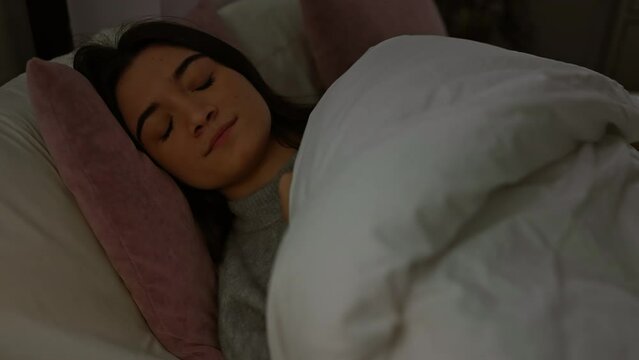 A young hispanic woman rests in bed, showing calm, comfortable, and smiling expressions in a cozy bedroom setting at night.