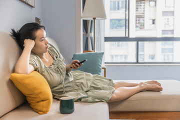 An Asian woman is watching TV in the living room at home, holding a TV remote control