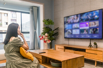 An Asian woman is watching TV in the living room at home, holding a TV remote control