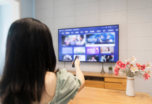 An Asian woman is watching TV in the living room at home, holding a TV remote control