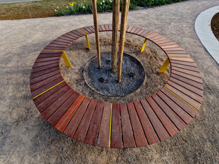 round bench made of wood encloses a tree, sitting around with wood paneling. metal frame. segments made up of planks around the tree. ventilation and watering grids at the roots