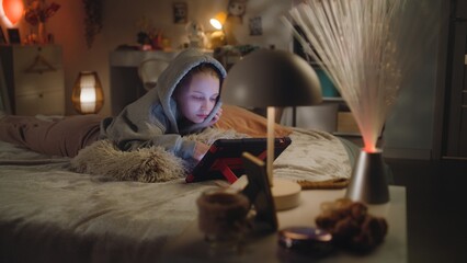 Young girl rests and lies on cozy bed in big comfort room in evening. Caucasian teen uses digital tablet computer to watch videos and surf Internet. Attractive teenager spends time in bedroom at home.