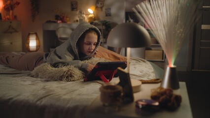 Young girl rests and lies on cozy bed in big comfort room in evening. Caucasian teen uses digital...