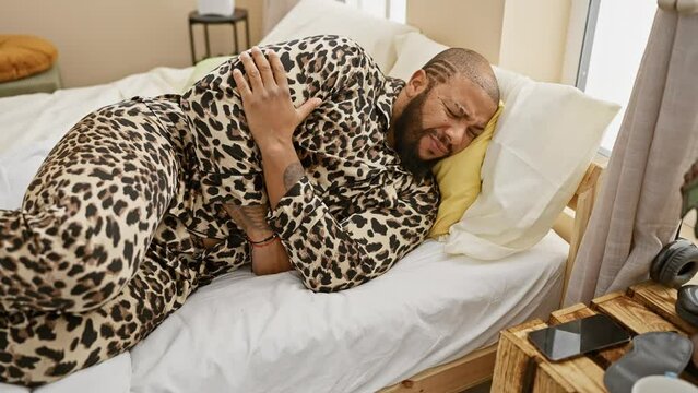 A bearded man in a leopard print pajama winces in pain, laying in a bedroom with a white bedspread and modern decor.