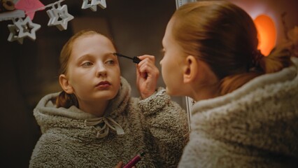 Young Caucasian girl does eyebrows makeup standing and looking in the mirror in cozy and comfort bedroom. Teenage girl spending leisure time and having fun at home at night. Concept of lifestyle. - obrazy, fototapety, plakaty