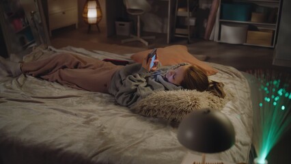 Caucasian teen chats with friends in social media and surfs the Internet using her mobile phone. Young girl rests and lies on big cozy bed in her room. Teenager spends time in comfort bedroom at home.