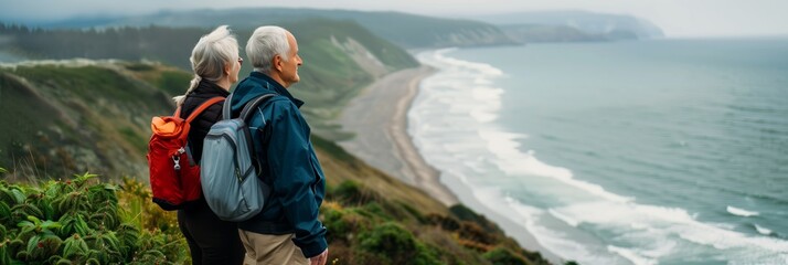 Elderly senior smiling active couple with backpacks enjoying hiking and walking along coastline near mountains and sea or ocean, active retirement lifestyle filled with outdoor adventures. - obrazy, fototapety, plakaty