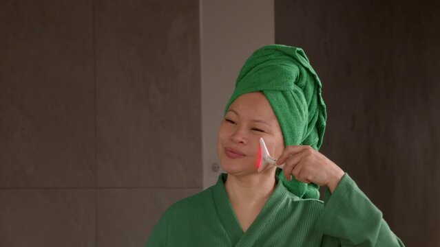 Charming middle aged Asian woman in dressing gown standing in front of mirror, making anti-aging beauty treatment using microcurrent facial massager for skincare and skin lifting in bathroom.