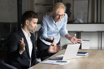 Two excited worried younger and elder business colleagues using laptop at workplace, staring at screen in surprise, speaking getting email, notice, message with unexpected concerning news