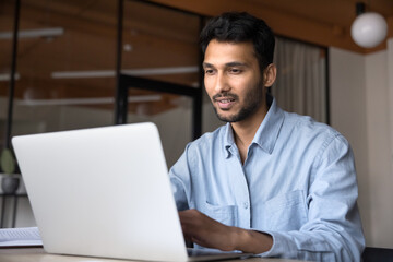 Positive young Indian business professional man in casual working at laptop in office, sitting at...