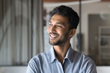 Cheerful young Indian man looking at window away with toothy smile, thinking on job success, successful career, creative work tasks, dreaming on future startup and profit. Male casual portrait - obrazy, fototapety, plakaty