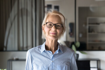 Positive blonde elder businesswoman in stylish glasses standing in office, looking at camera. Happy confident senior female manager, entrepreneur, business coach woman head shot portrait