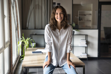Cheerful young entrepreneur woman in casual clothes posing in office, leaning on workplace table in modern workspace, looking at camera, smiling, laughing, enjoying, enjoying successful career - obrazy, fototapety, plakaty