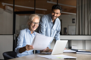 Obraz premium Two cheerful successful diverse business colleagues reviewing paper financial reports, reading legal documents, agreement at laptop, doing paperwork together, smiling, laughing,