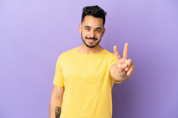 Young caucasian man isolated on purple background smiling and showing victory sign