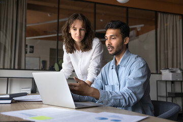 Two serious young Indian and Caucasian project managers working at laptop together, discussing online content, pointing hand at display, speaking, sitting and standing at workplace table