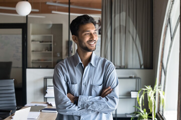 Cheerful handsome young Indian manager man in casual shirt standing in office with hands crossed, looking away, smiling, thinking on business ideas, successful marketing strategy - obrazy, fototapety, plakaty